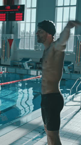 Medium full vertical shot of muscular Caucasian male swimmer in cap and trunks standing by pool, doing warmup exercises, rotating shoulders, flexing, stretching, swinging arms to prepare for practice
