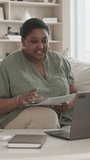 Vertical shot of black woman sitting of sofa in living room and discussing documents via online video call on laptop during e-meeting