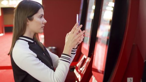 Teenager buying movie ticket from vending machine at movie theater at mall. Woman using mobile to read information about movies online. Making photos of screen, downloading information from internet