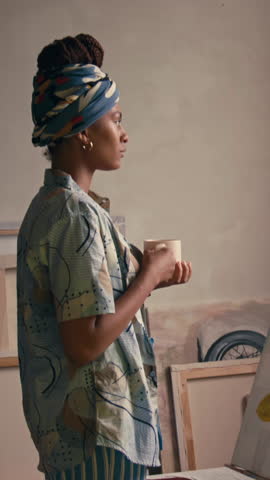 Vertical handheld side footage of young Black fashionable artist in bandana drinking tea or coffee while thinking in art studio and looking out of window