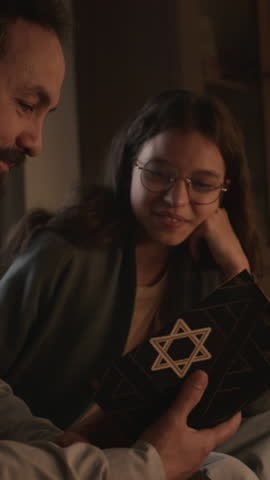 Close-up vertical shot of face and hands of middle-aged religious Jewish man and teenage girl sitting together at home, reading Torah with star of David on cover, and discussing commandments