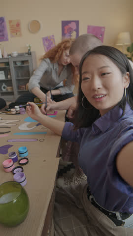 Vertical handheld POV of young Asian female activist recording video blog of herself with other diverse feminist women drawing banners and chatting while getting ready for protest together at home