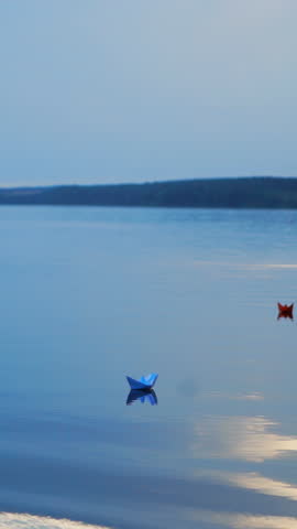 Colored paper ships go far away from the shore on the blue water background at sunset. Homemade boats with different colors floating on the big pond