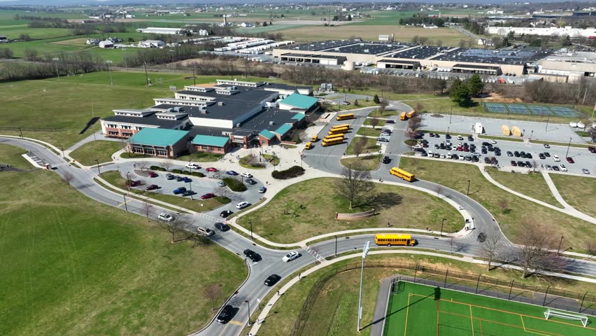 Drone flight over american school with parking yellow buses and traffic on roundabout. High School during sunny day in rural scenic area. Aerial descending top down shot.