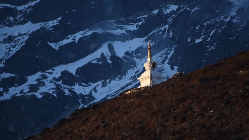 Close up view of a sun lit white stupa in front of a massif towering montain slope in the Himalayas.