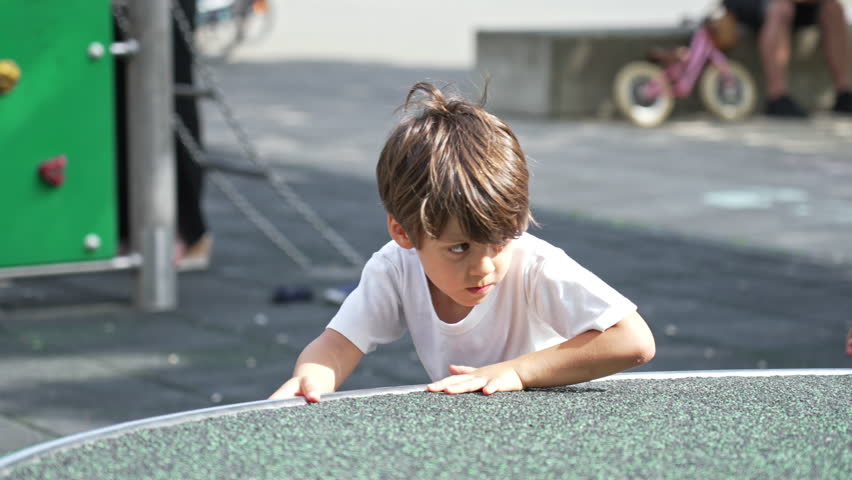Child Spins Merry-Go-Round At Playground Park During Sunny Day. Young Boy Engaged In Play Demonstrating Strength And Speed