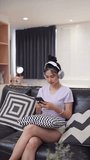 The Asian woman is choosing and listening to music on her smartphone to relax and pass the time while sitting on the sofa in her home.
