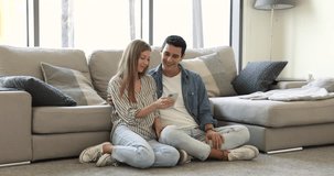 Smiling Hispanic loving couple resting seated on floor in spacious living room with modern smart phone, enjoy online communication, making order, using delivery e-services, spend pastime on internet