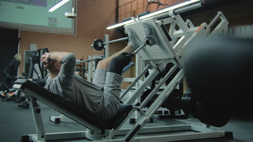Motivated athlete with prosthetic leg trains on leg press machine in modern fitness center. Adult sporty man with artificial limb performs strength exercises using professional sports gym equipment.