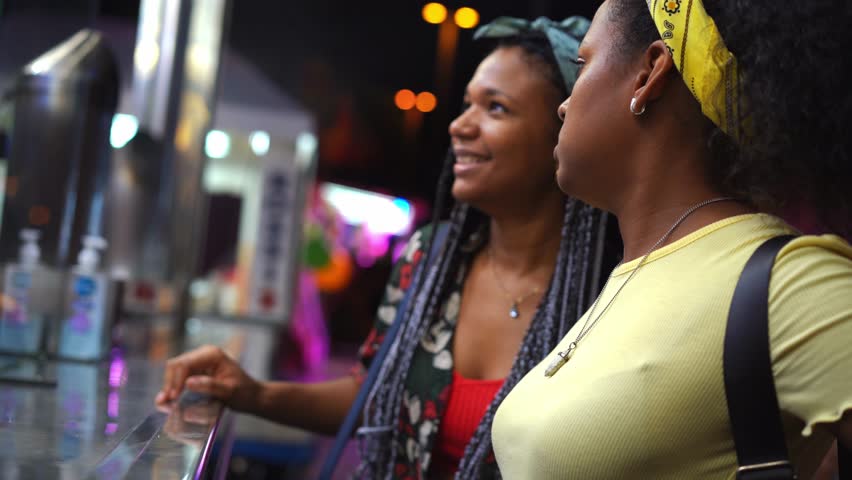 Two happy young women buying fritters at fast food stand at outdoor amusement park. Lesbian girl couple in love enjoying a date. Happy and friendly African American people. 