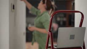 In the foreground is an open laptop lying on the step of a red stepladder. Out of focus, a woman in a green overalls paints the corridor wall with a paint roller