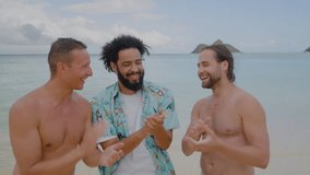 Group of three tourists, African American man in Hawaiian shirt, two shirtless Caucasian guys clapping hands, laughing, standing on beach near the ocean. High quality 4k footage - Powered by Shutterstock - Get 15% off with code: PIKWIZARD15