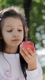 Preschool little girl eating red apple. Kid eating fruit. Child bites fruit and chews looking at camera in park. Healthy diet - vegetarian. Vertical video