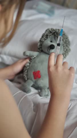 Vertical screen: a little girl uses a syringe with a needle to inject her plush toy into piercing the paw of a hedgehog. An 8 year old child plays with soft toys in a veterinary hospital in bed. Royalty-Free Stock Footage #3466311833