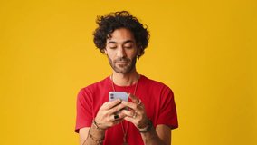 Man with curly hair, dressed in red T-shirt, using mobile phone, typing massages isolated on yellow background in studio