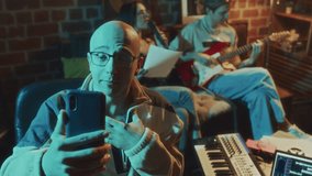 Panning footage of Caucasian bald blogger filming vlog on phone while his friends singing song to guitar on sofa in background in vintage cozy studio