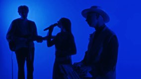 Side footage of silhouettes of rock band of keyboarder, guitarist and singer performing music in obscure professional studio with blue neon filter
