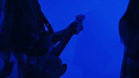 Side tilt closeup of male guitarist playing electric bass guitar and singing while performing music with band in blue neon lit studio
