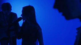 Side footage of silhouettes of female singer and musicians performing music in professional studio with blue colored filter
