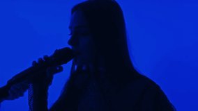 Side closeup of silhouette of female singer with microphone performing song with music band in studio with blue colored filter