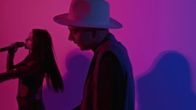 Side tilt footage of male keyboard player in hat accompanying female singer on synthesizer while performing music in studio with pink colored neon filter