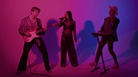 Full footage of charismatic Caucasian glam rock band performing song accompanying on bass guitar and synthesizer in studio with pink and purple colored neon filter