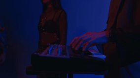 Side tilt footage of male keyboardist playing on synthesizer during rock concert with music band in blue neon lit studio or club