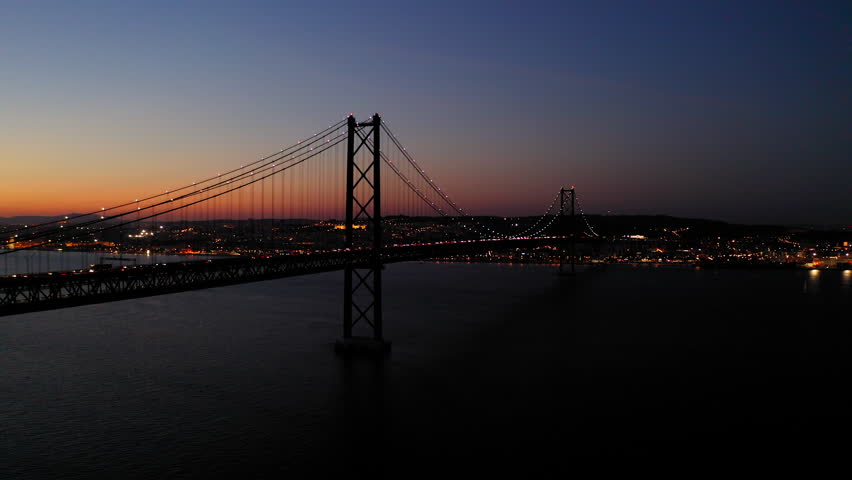 Aerial Panning Shot Of Illuminated Famous Suspension Bridge In City, Drone Flying Over Tagus River During Scenic Night - Lisbon, Portugal