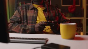 Waist up of young African American male gamer wearing checkered shirt, cap and headset with mic getting upset after losing game, sitting with controller at desk in neon light room at night