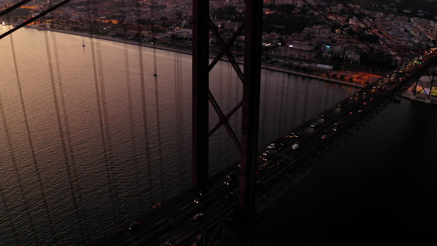 Aerial Tilt Up Backward Shot Of Illuminated Cars Moving On Famous Bridge Over Tagus River Against Orange Sky At Dusk - Lisbon, Portugal