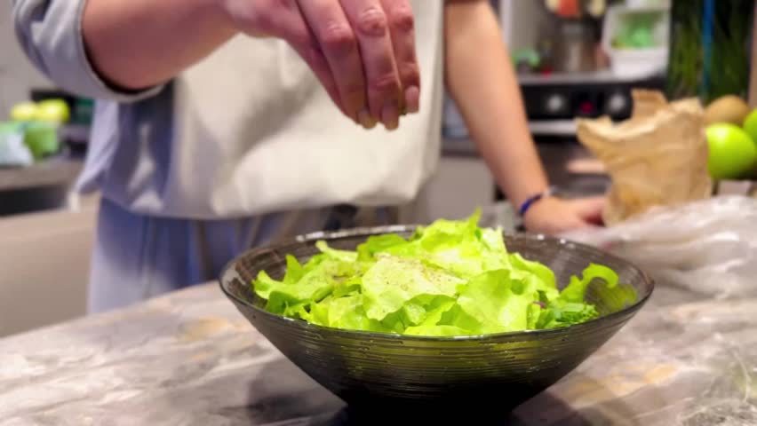 A person sprinkling salt on a bowl of lettuce. The person is wearing a white shirt