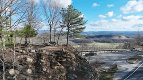 A panning video of a scenic overlook. The shot includes a stand of trees on a rocky outcropping with rolling hills and mountains in the background. The shot pans over to a winding road.