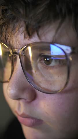 A vertical portrait of a teenage boy in large glasses chatting on the Internet in front of the monitor at night. Smart 11 year old boy in an online game. Online education remotely for children. Royalty-Free Stock Footage #3466750847