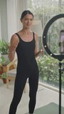 Vertical shot of female fitness influencer talking in front of smartphone on tripod with ring light while filming vlog or going live on social media at home