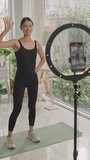 Vertical shot of female fitness blogger doing jumping jacks in front of smartphone on tripod with ring light while filming video workout for social media at home