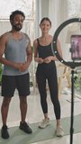 Vertical shot of male and female fitness bloggers standing in front of smartphone on tripod with ring light and communicating with subscribers during live stream on social media