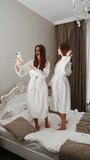 Two young women in bathrobes dance on the bed and film themselves. Hotel holiday concept. Vertical video