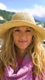 Portrait of happy attractive young woman wearing straw hat looking and smiling at the camera with seacoast and yachts background. A vertical video.