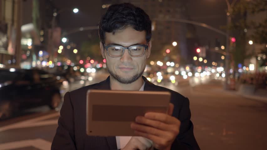 Businessman Focus on Touchpad against the Backdrop of Illuminated Skyscrapers
