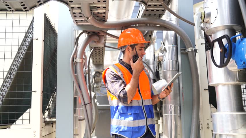 caucasian male engineer wearing a reflective jacket and safety helmet walks with a tablet to inspect the progress of electrical piping work on the roof of a building.
