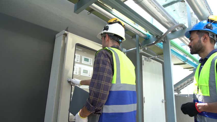 Caucasian male engineer and construction contractor wearing reflective jackets and helmets for safety walk with tablets inspecting electrical control cabinets on the rooftop of a building.