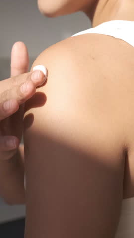 Woman applying skin care cream. Happy smiling young female applying sunscreen or moisturizer lotion on a shoulder. Beauty skin concept. Body care at home interior on a sunny day. Close up