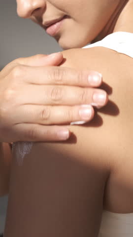 Woman applying skin care cream. Happy smiling young female applying sunscreen or moisturizer lotion on a shoulder. Beauty skin concept. Body care at home interior on a sunny day. Close up