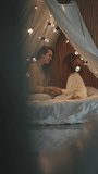 Cute mum with her daughter kid child sitting in a cozy tent with lights, bonding embracing hugging. Happy childhood with mother.