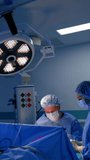 Caucasian male surgeon works at operation assisted by female scrub nurse. Doctor sits at the patient under the round lamp hanging on the ceiling. Vertical video.