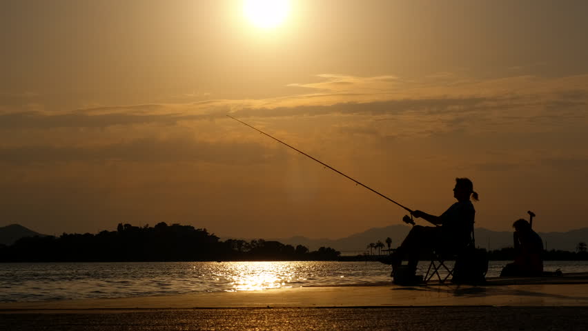 Woman fishing by sunset city pond. A woman silhouette sitting in the chair and fishing on the evening beach by waterfront in summer. A concept of summer hobby by the sea.