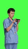Side view Medical assistant playing mobile video games on smartphone app, taking a break from healthcare work. Nurse enjoys gaming tournament on phone, standing against greenscreen backdrop. Camera B.