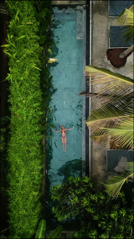 Aerial view of a serene woman floating leisurely in a secluded swimming pool, surrounded by lush tropical flora, capturing the essence of tranquility and holiday relaxation at an exotic destination.