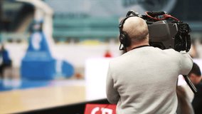 Back view on cameraman working on professional basketball sport event match. Camera operator following puck in dynamic in sport game. Concept of technology, action.