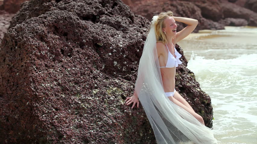 Attractive bride with long veil leans on rock enjoying gentle waves. Blonde woman in white bikini laughs posing during weeding photoshoot